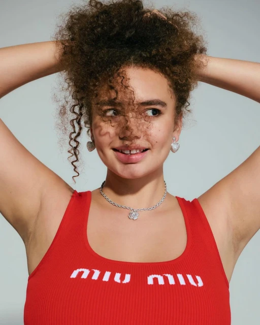 Portrait of a young adult woman with curly brown hair, wearing a red top and jewelry, smiling and looking off to the side on a neutral backdrop.