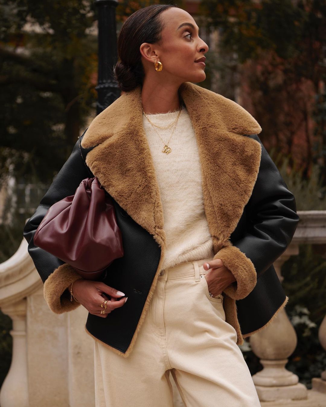 Portrait of a woman wearing a black shearling jacket, off-white sweater, light-wash jeans and holding a dark red leather handbag.