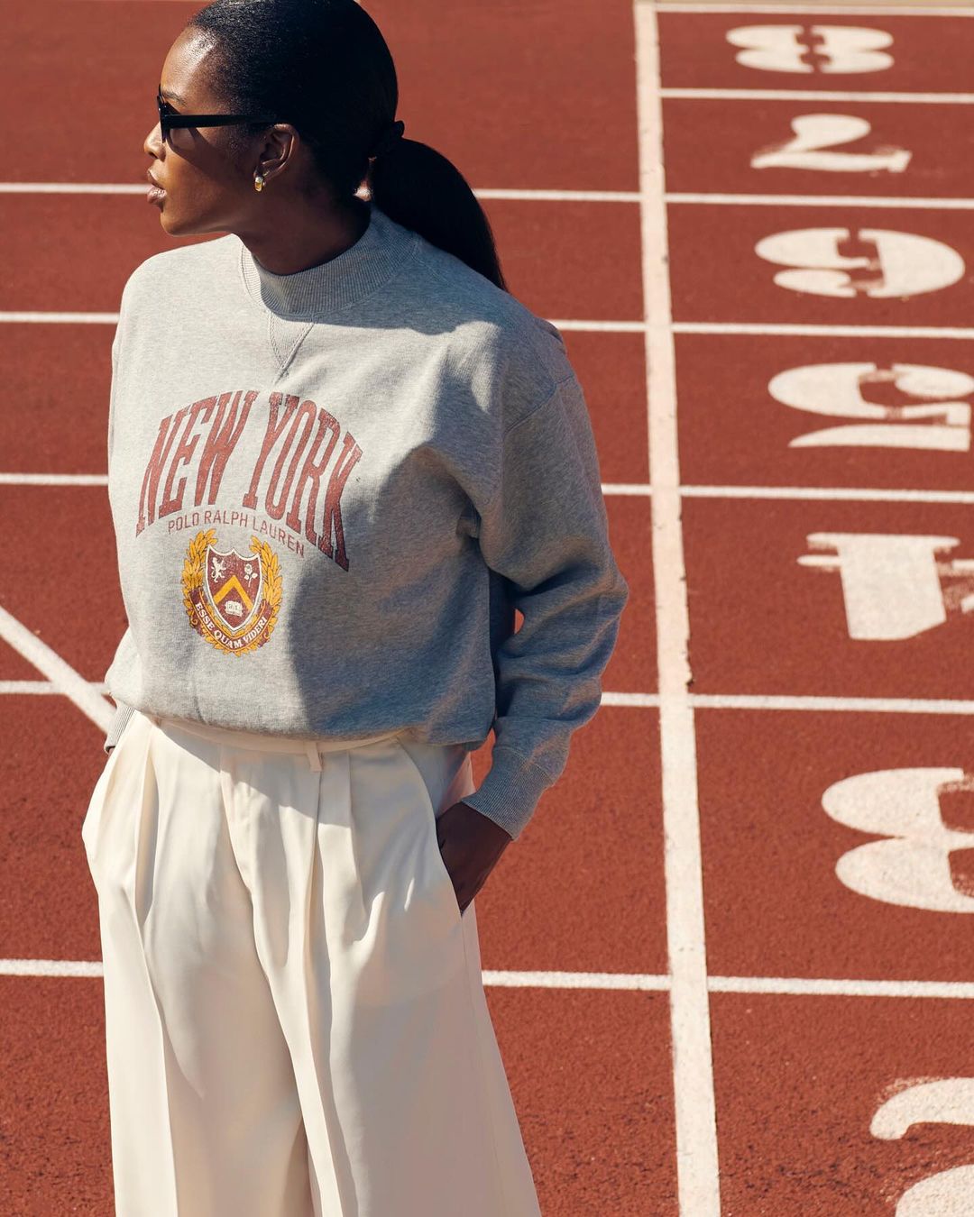 Woman in grey 'New York' sweatshirt and white pants stands on a red running track, posing in fashion shot with athletic backdrop.