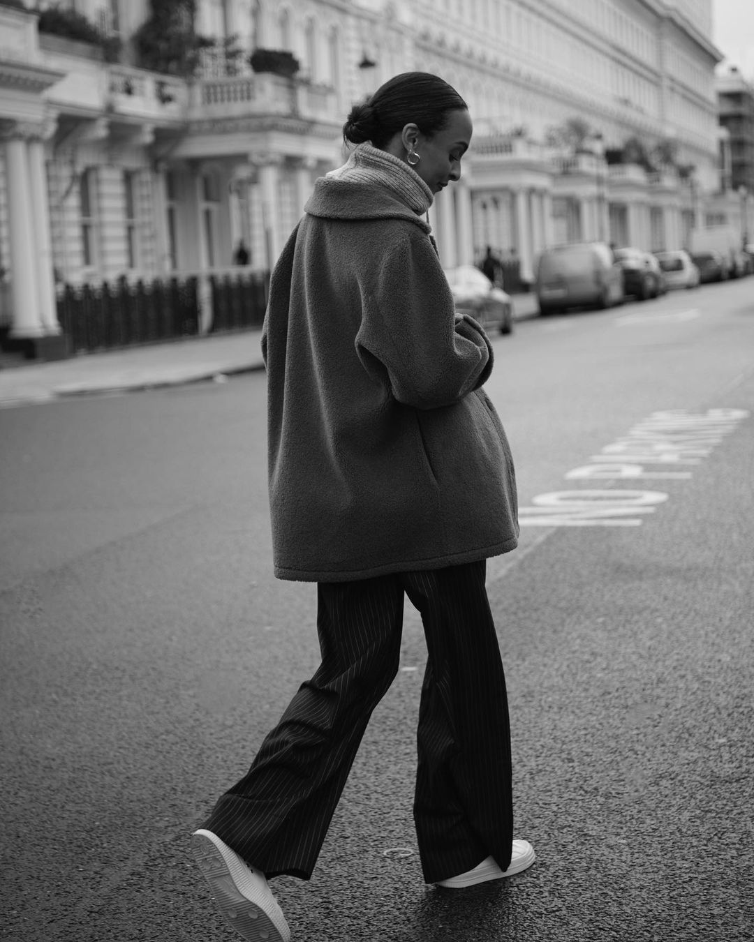 Monochrome portrait of an adult woman in a coat crossing a city street with row houses in the background, captured at eye-level.