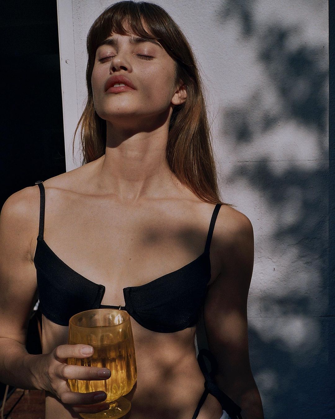 Woman in black bikini top and bottom poses outdoors in dappled sunlight while holding yellow glass, eyes closed and serene expression.
