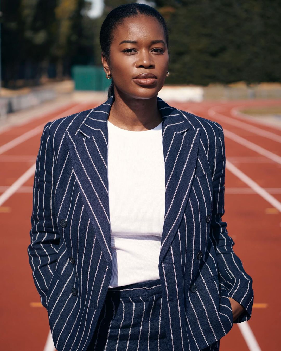 A confident African American woman in a pinstripe suit stands on a red running track with green foliage in the background, in natural light.