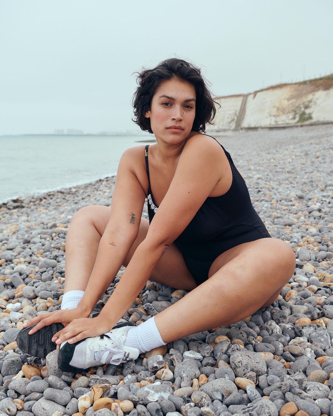 Woman in a black swimsuit sits on a pebble beach wearing socks and sneakers; rocky shore, cliffs, and overcast sky in the background.