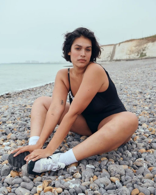 Woman in a black swimsuit sits on a pebble beach wearing socks and sneakers; rocky shore, cliffs, and overcast sky in the background.