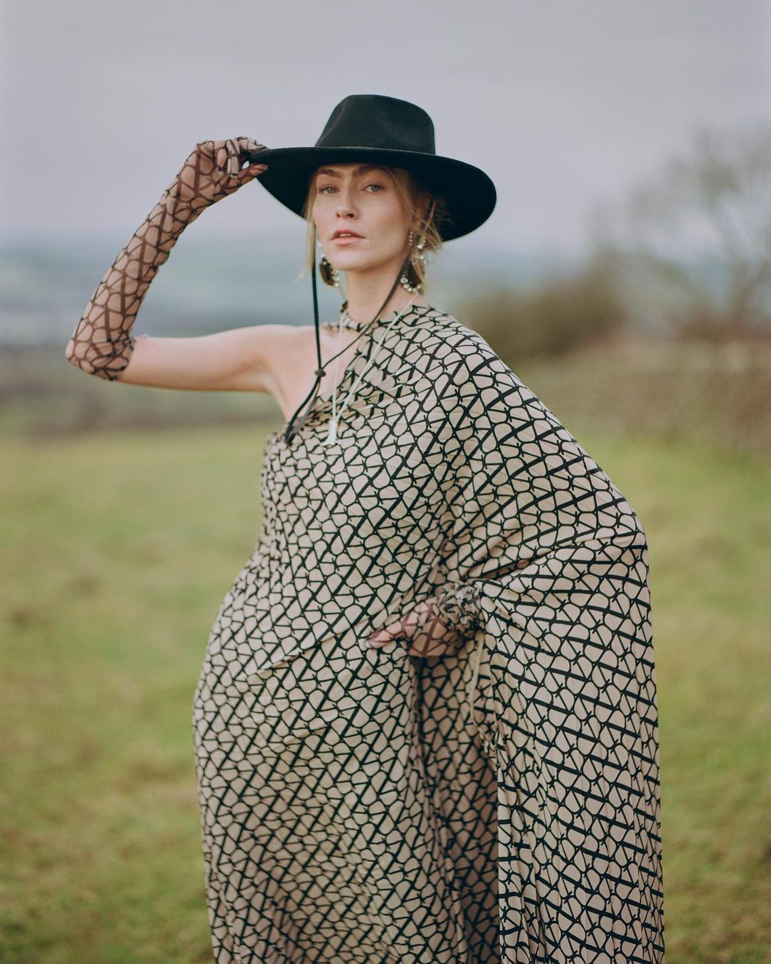 Adult female wearing patterned dress and cowboy hat poses in a field on a cloudy day in a medium shot with shallow depth of field.
