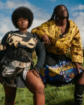 Two dark-skinned young women pose in a grassy field against a blue sky. One has a large afro and is wearing a patterned top and skirt.