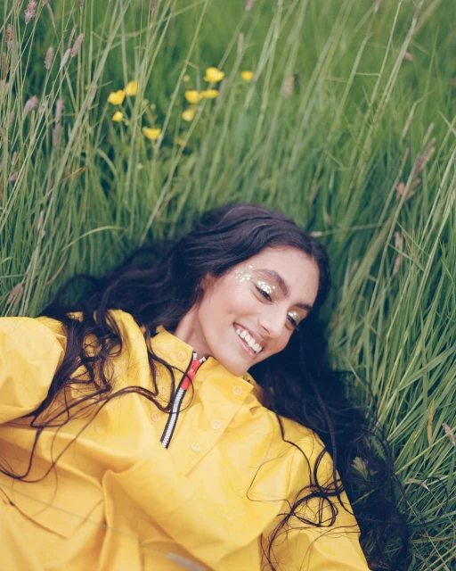 Smiling woman in a yellow raincoat with glitter makeup lying in a field of tall grass and yellow flowers in a medium outdoor shot.
