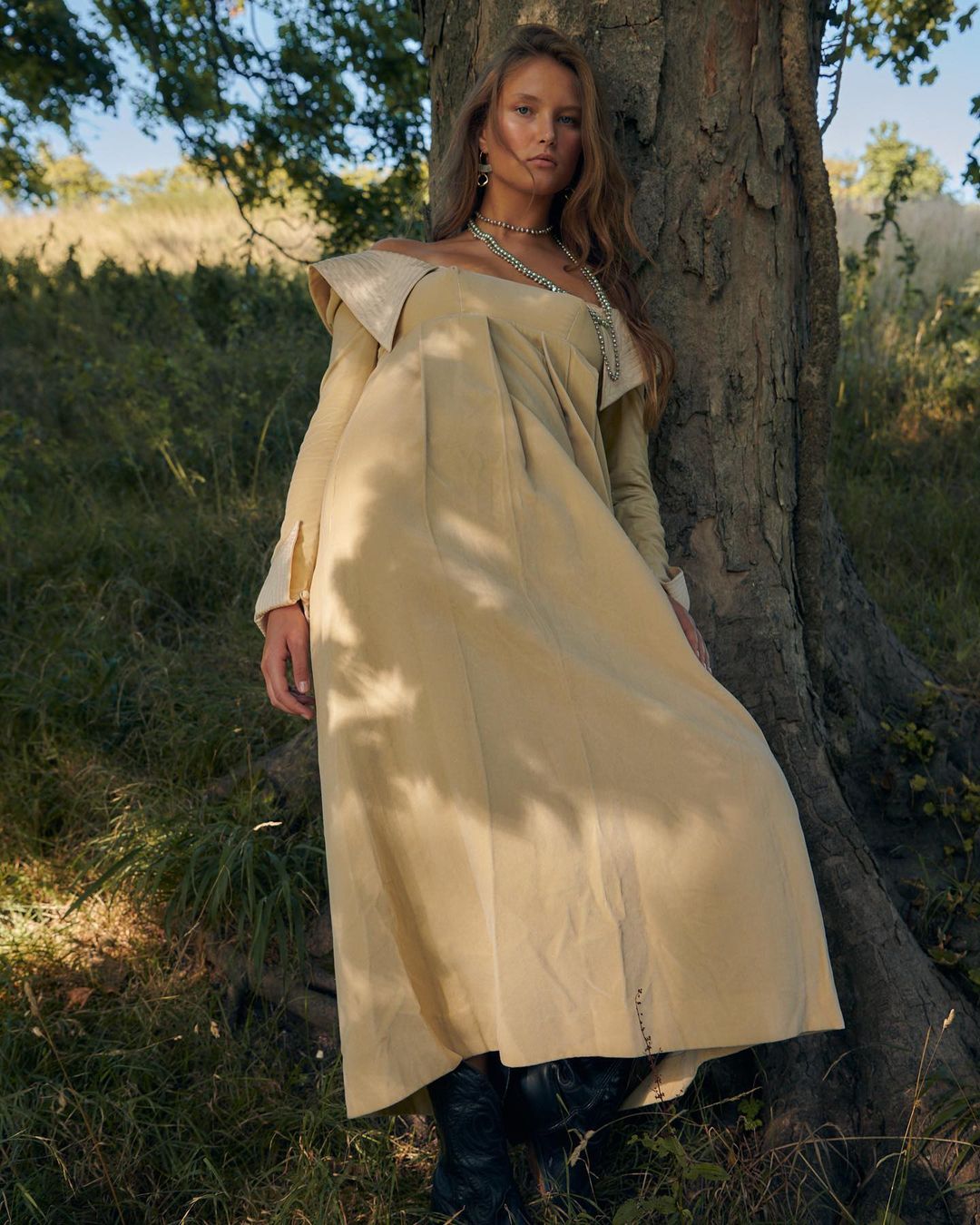 A woman with long brown hair stands next to a tree in a light-beige, off-the-shoulder dress with long sleeves, in a grassy outdoor setting.