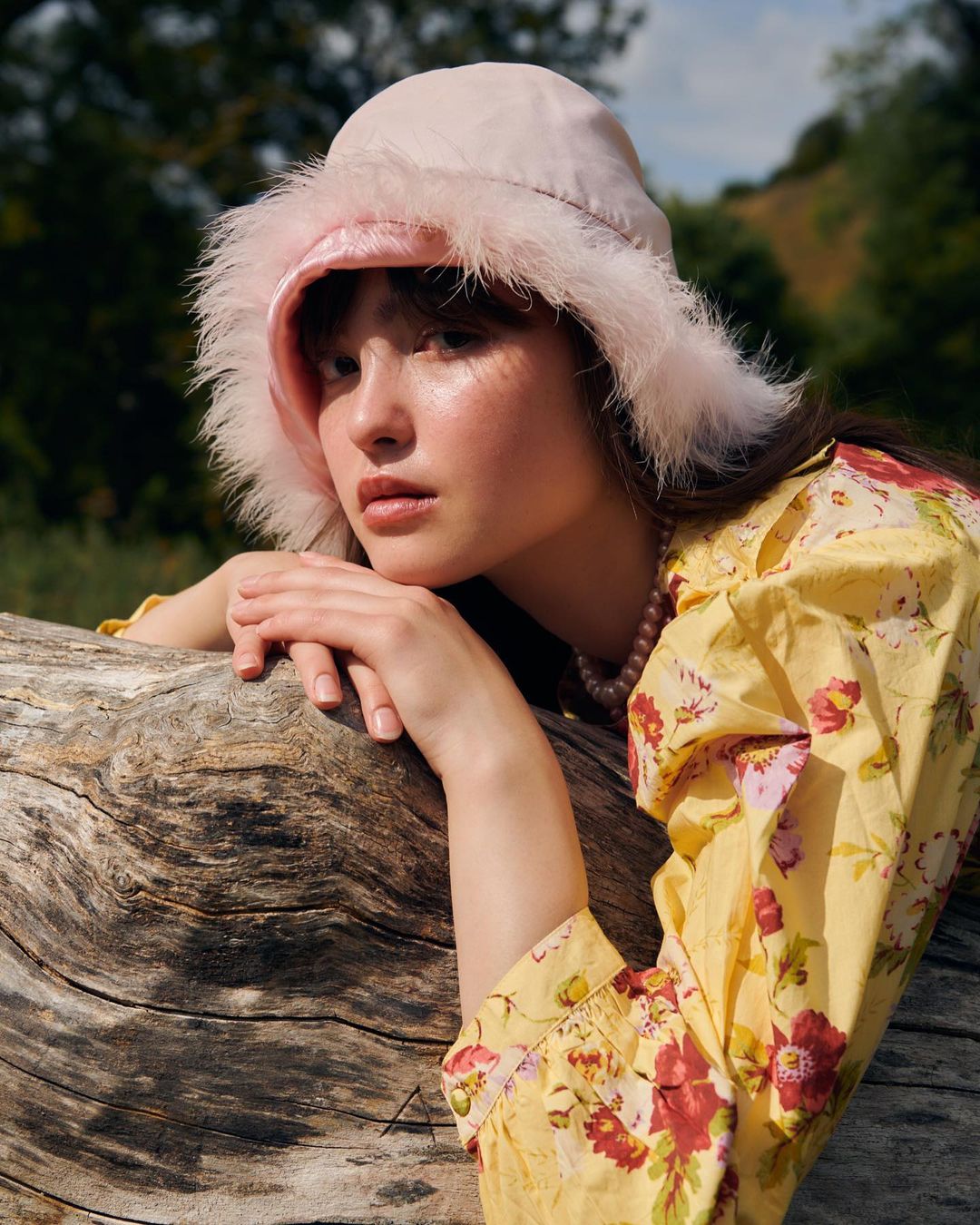 Young woman in a floral blouse poses outdoors wearing a pink fur-trimmed bucket hat while resting her arms on a fallen tree trunk.