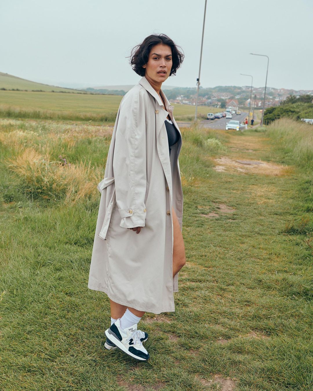 A woman with dark hair in a trench coat poses on a grassy path, wearing sneakers and white socks with a field and sky behind her.