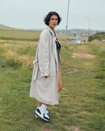 A woman with dark hair in a trench coat poses on a grassy path, wearing sneakers and white socks with a field and sky behind her.