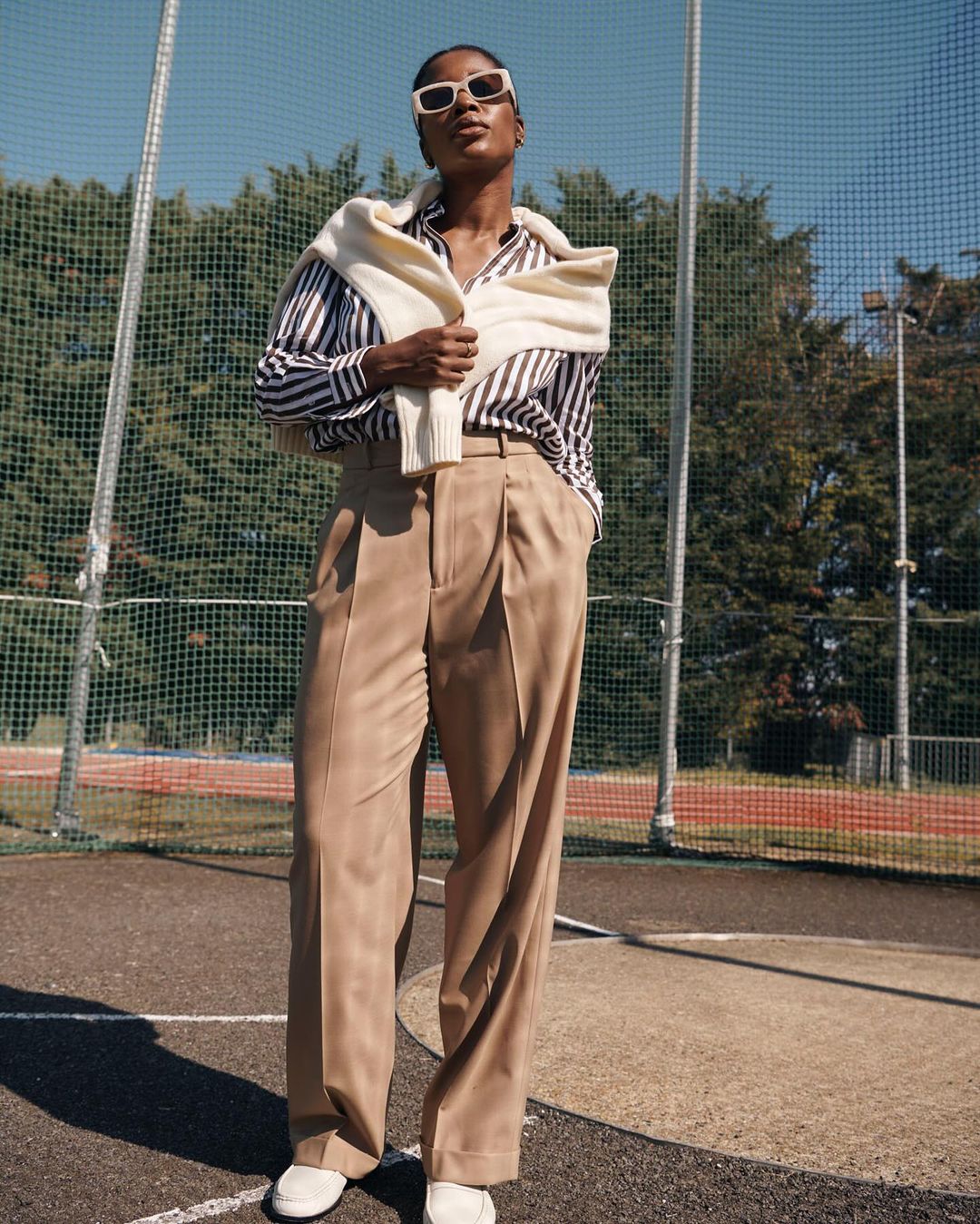 A fashionable Black woman poses in a striped shirt, wide-legged trousers, white shoes and sunglasses, with a sweater around her shoulders.