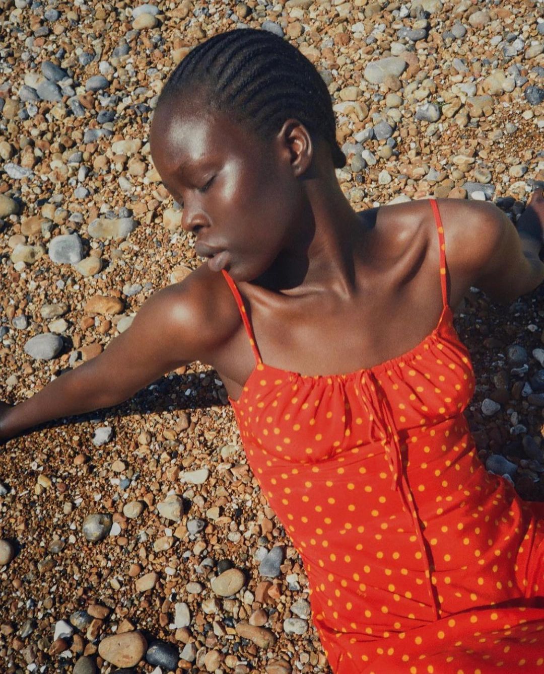 Woman in red and yellow polka dot dress lies on a pebble beach, eyes closed in sunlight, showcasing fashion and beauty.
