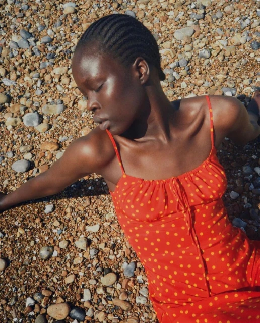 Woman in red and yellow polka dot dress lies on a pebble beach, eyes closed in sunlight, showcasing fashion and beauty.