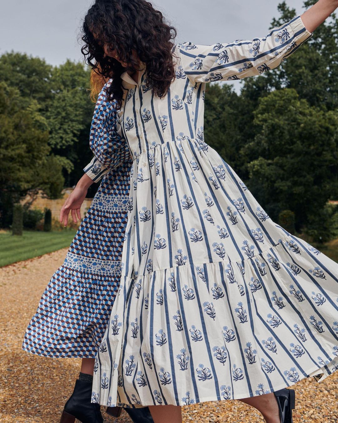 Two adult women in patterned dresses walking on a gravel path outdoors, with trees and greenery visible in the background.