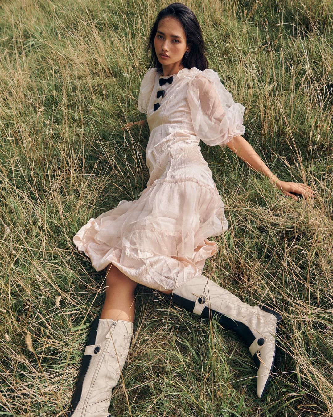 Young woman in a light pink dress with knee-high boots lying in tall grass in an outdoor setting for a fashion editorial shoot.
