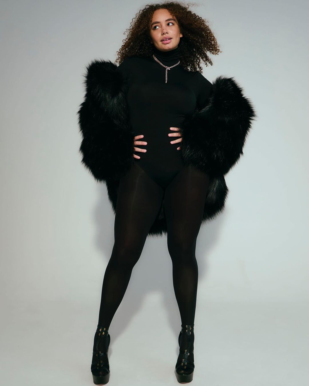 Model with curly hair wearing a black turtleneck bodysuit, black tights, and a fluffy black faux fur coat against a white background