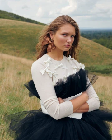 Model with brown hair wearing a cream sweater with white bows and a black tulle skirt outdoors in a grassy meadow.