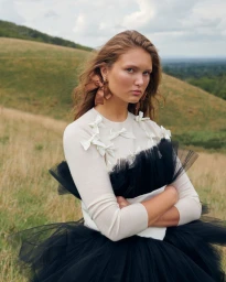 Model with brown hair wearing a cream sweater with white bows and a black tulle skirt outdoors in a grassy meadow.