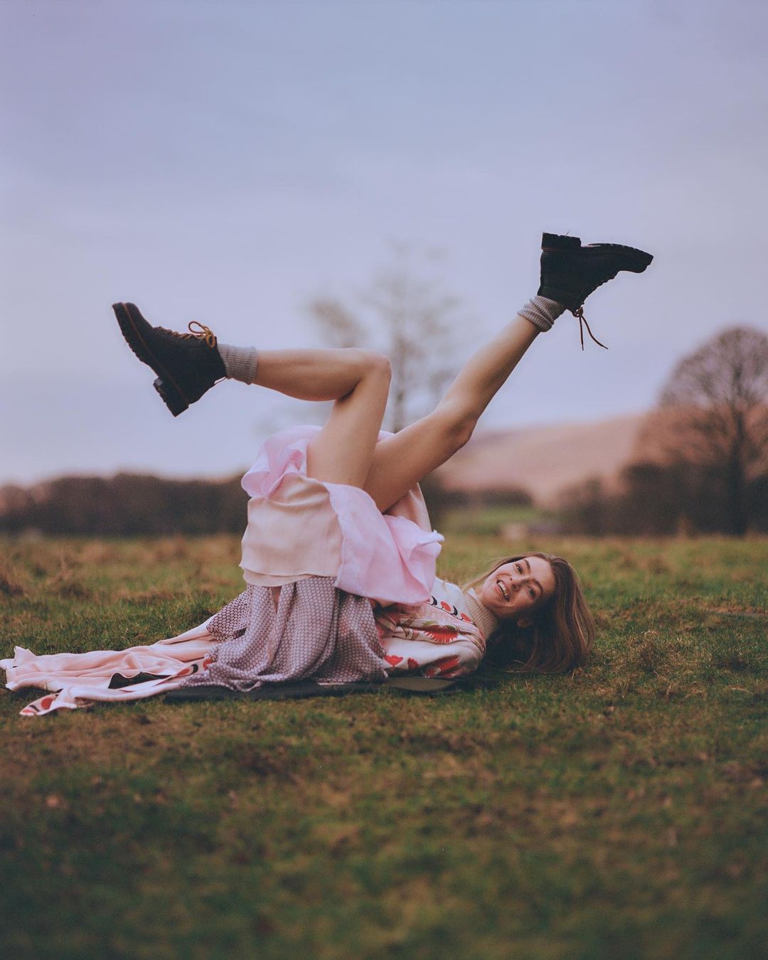 Woman lying on grassy field with legs in the air wearing a ruffled skirt and black boots