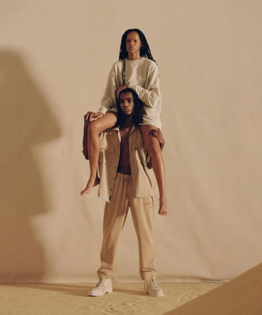 A man in beige pants carries a woman on his shoulders in a studio setting, both wearing matching outfits against a neutral backdrop.
