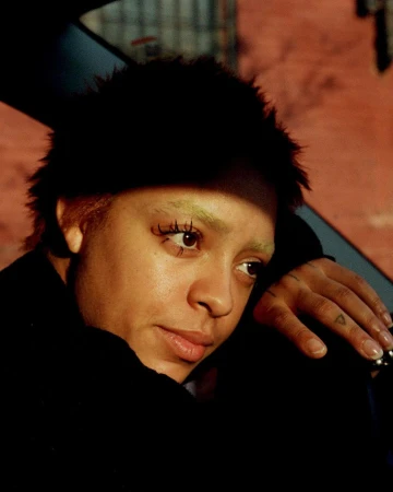 Close-up portrait of a woman with short dark hair and light eyebrows resting her head on her hand, photographed by Clifford Prince King.