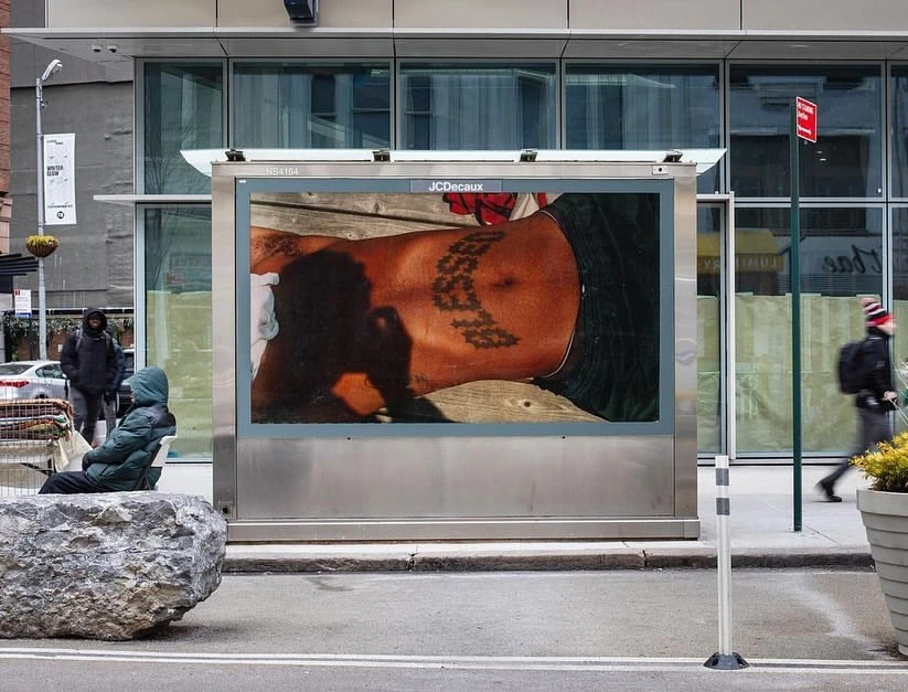 An advertisement on a bus stop shelter features a tattooed torso; people walk by in an urban street setting with a person sitting nearby.