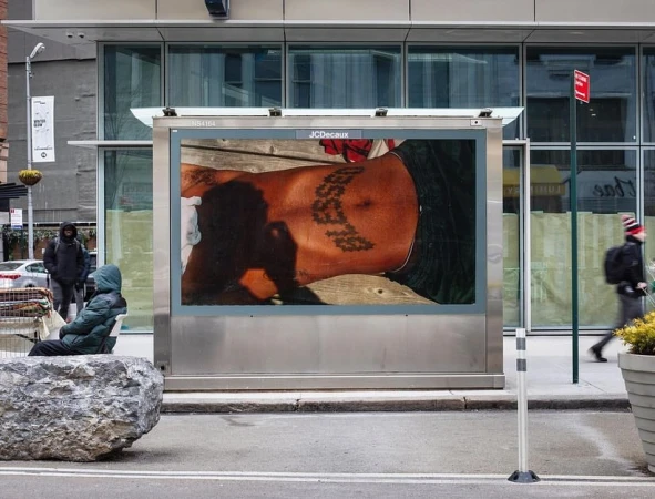 An advertisement on a bus stop shelter features a tattooed torso; people walk by in an urban street setting with a person sitting nearby.