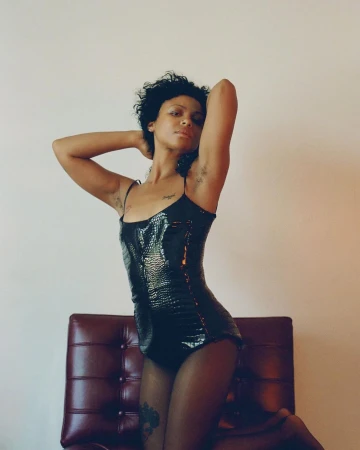 Black woman wearing a glossy black patterned leotard posing against a plain wall behind a dark red tufted leather chair