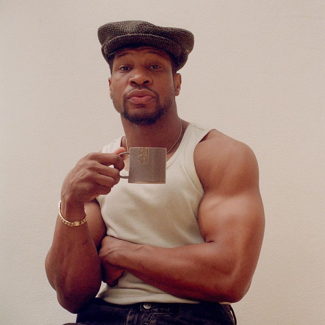 Adult male with muscular build wears a cap and tank top while holding a mug, posing indoors against a neutral background.