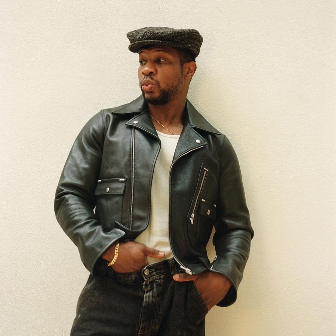 A stylish African-American man with a cap poses indoors, wearing a leather jacket, white shirt, and grey jeans, against a neutral backdrop.