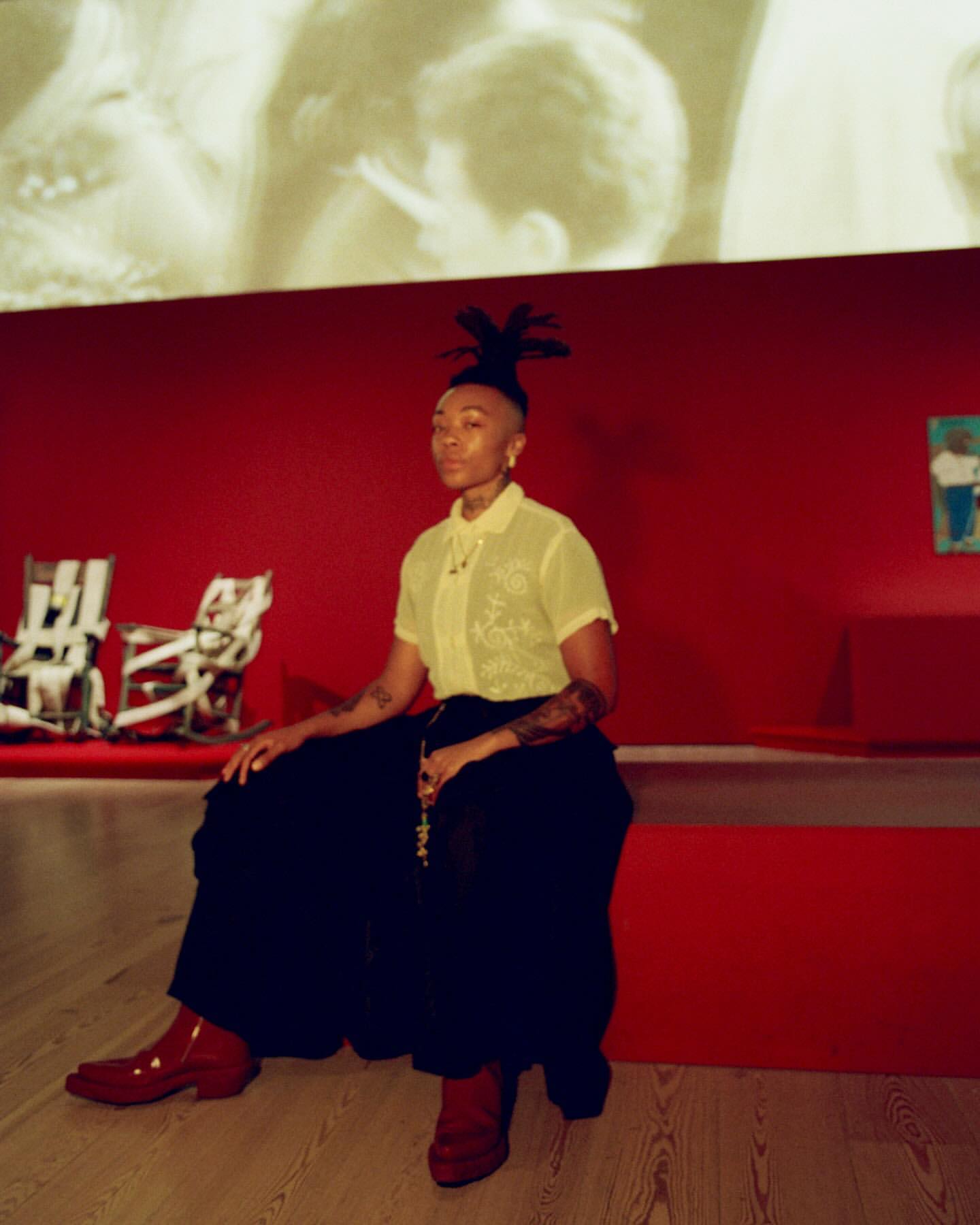 Adult female with dreadlocks and tattoos sitting on a red bench wearing a yellow collared shirt, black pants, and red platform boots indoors.