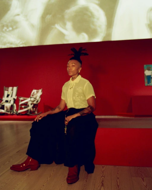 Adult female with dreadlocks and tattoos sitting on a red bench wearing a yellow collared shirt, black pants, and red platform boots indoors.