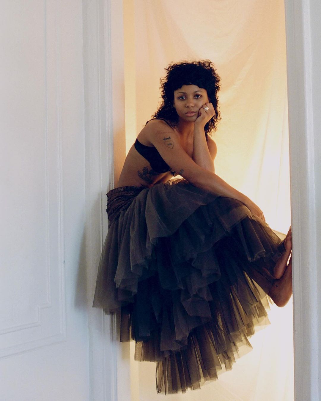 An adult woman with dark hair and a black bra sits on a white ledge wearing a large tiered tulle skirt in a light-filled indoor setting.