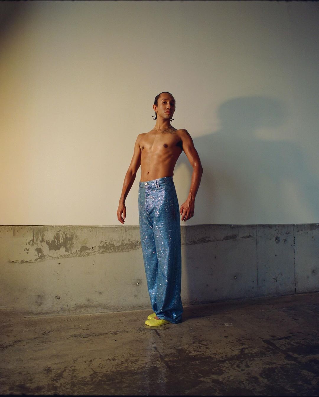 Adult male poses shirtless wearing shiny blue pants and yellow flats against a neutral wall and concrete base in an indoor setting.