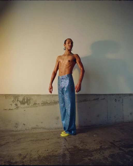 Adult male poses shirtless wearing shiny blue pants and yellow flats against a neutral wall and concrete base in an indoor setting.