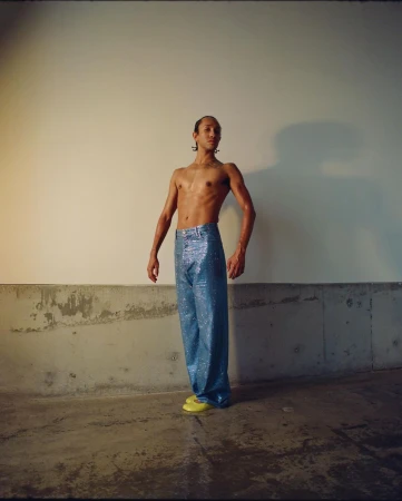 Adult male poses shirtless wearing shiny blue pants and yellow flats against a neutral wall and concrete base in an indoor setting.