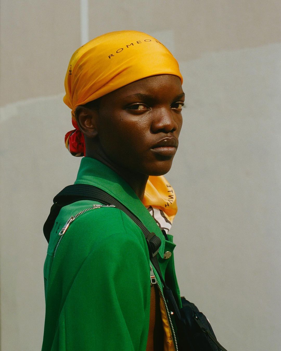 Portrait of an African male model wearing a yellow headscarf and a green jacket, looking at the camera against a neutral background.