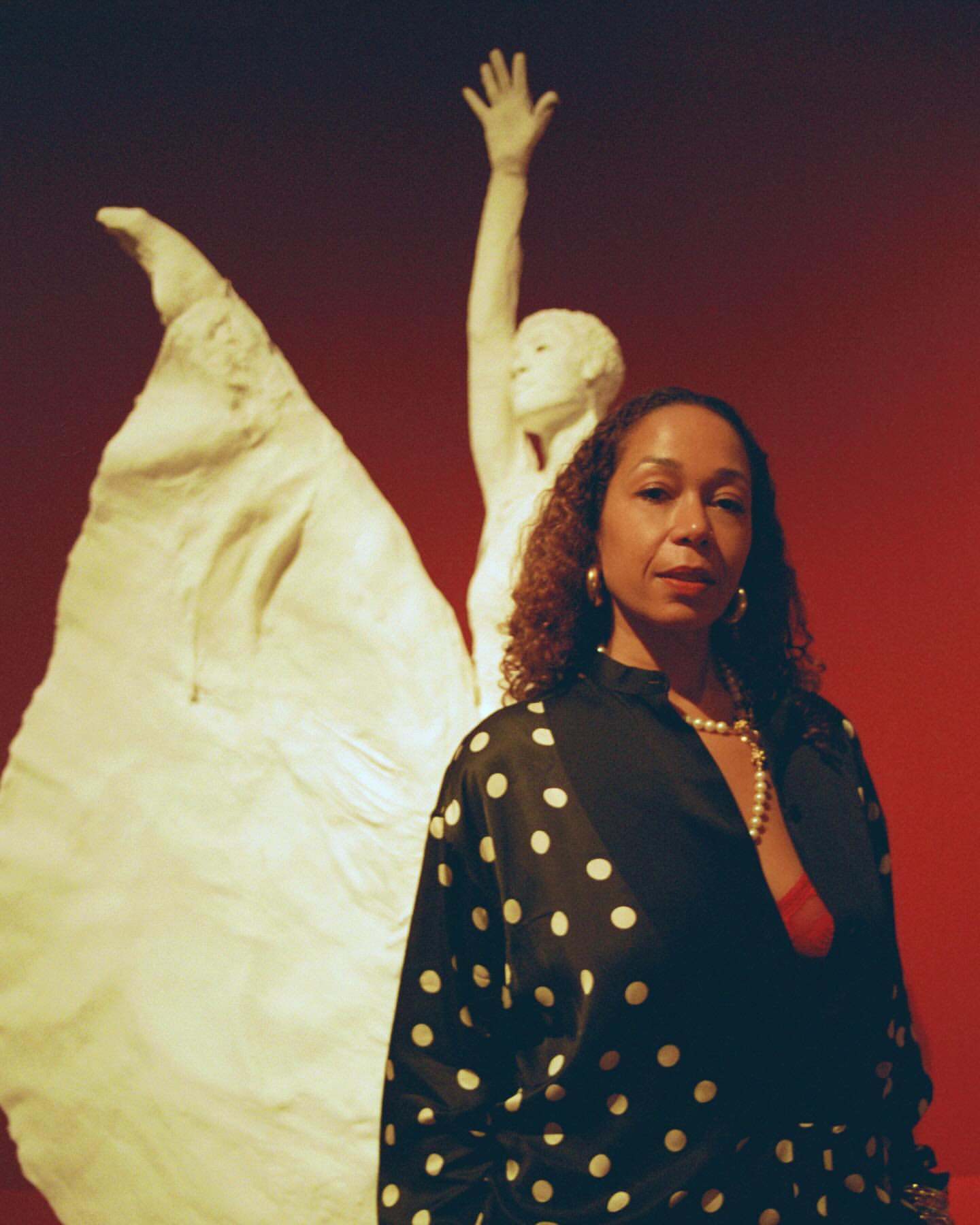 Adult woman poses in front of a light stone sculpture against a warm red background, wearing a black and white polka dot blouse.