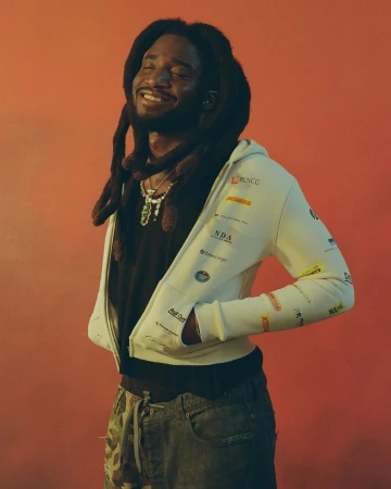 Portrait of a smiling Black man with long locs wearing a white zip-up hoodie against a warm orange wall.