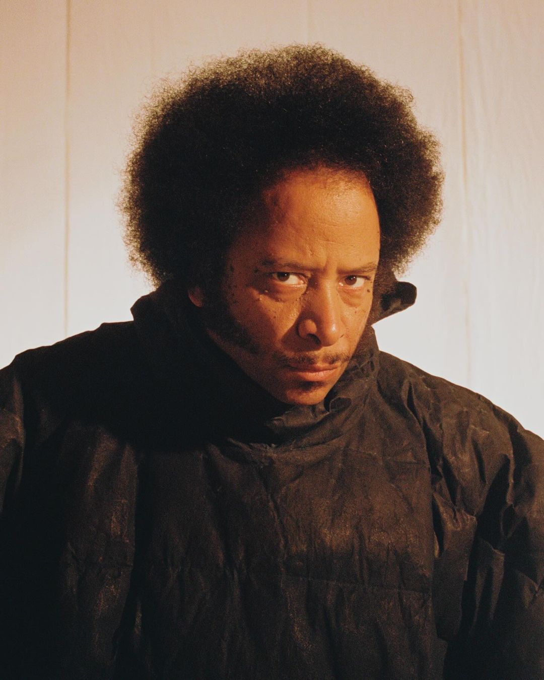 Portrait of an adult male with an afro, dark jacket, and a slight smirk, captured in a close-up shot against a gradient background.