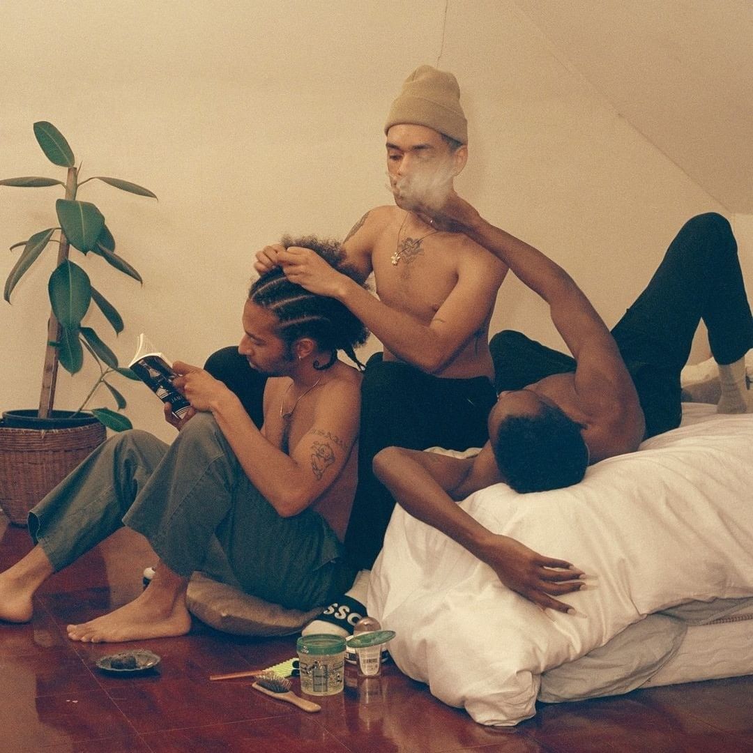 Three shirtless young men relax indoors; one reads a book, another has his hair styled, and a third reclines on a white bed.