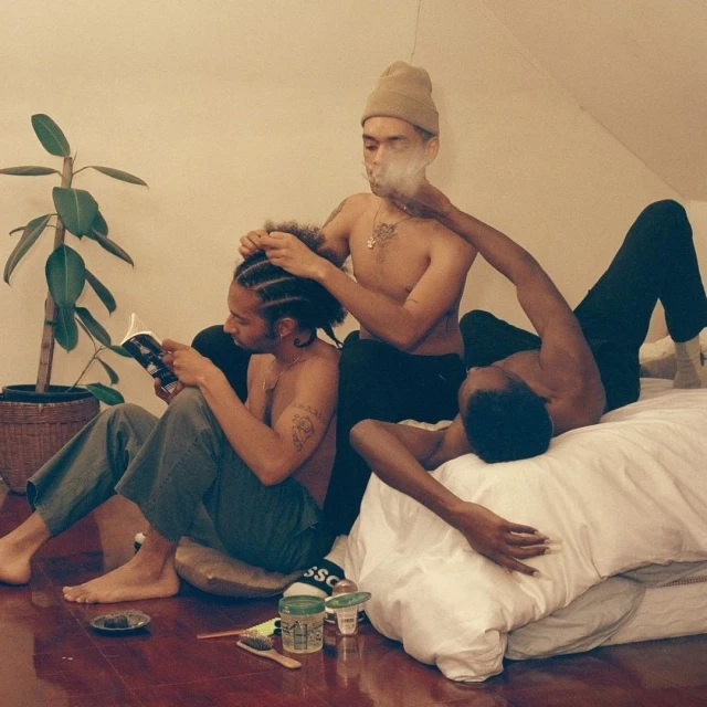 Three shirtless young men relax indoors; one reads a book, another has his hair styled, and a third reclines on a white bed.