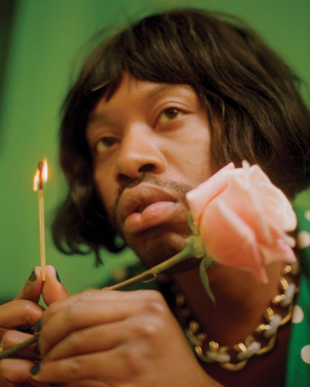 Portrait of a person with a short black wig holding a pale pink rose and a lit match against a solid green backdrop in indoor lighting.