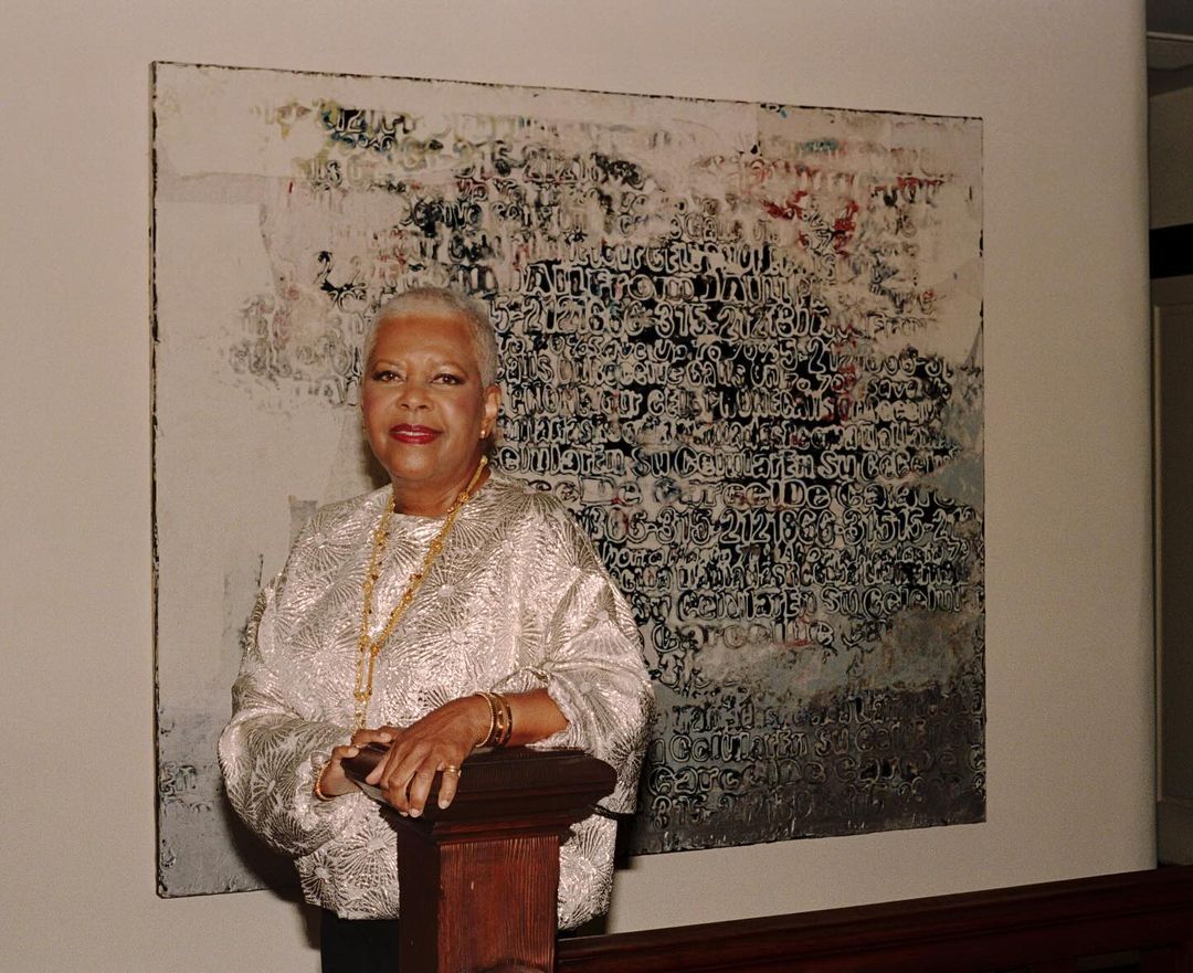 Elegant woman in a patterned jacket poses near an abstract painting. Gold jewelry and warm lighting enhance the sophisticated atmosphere.