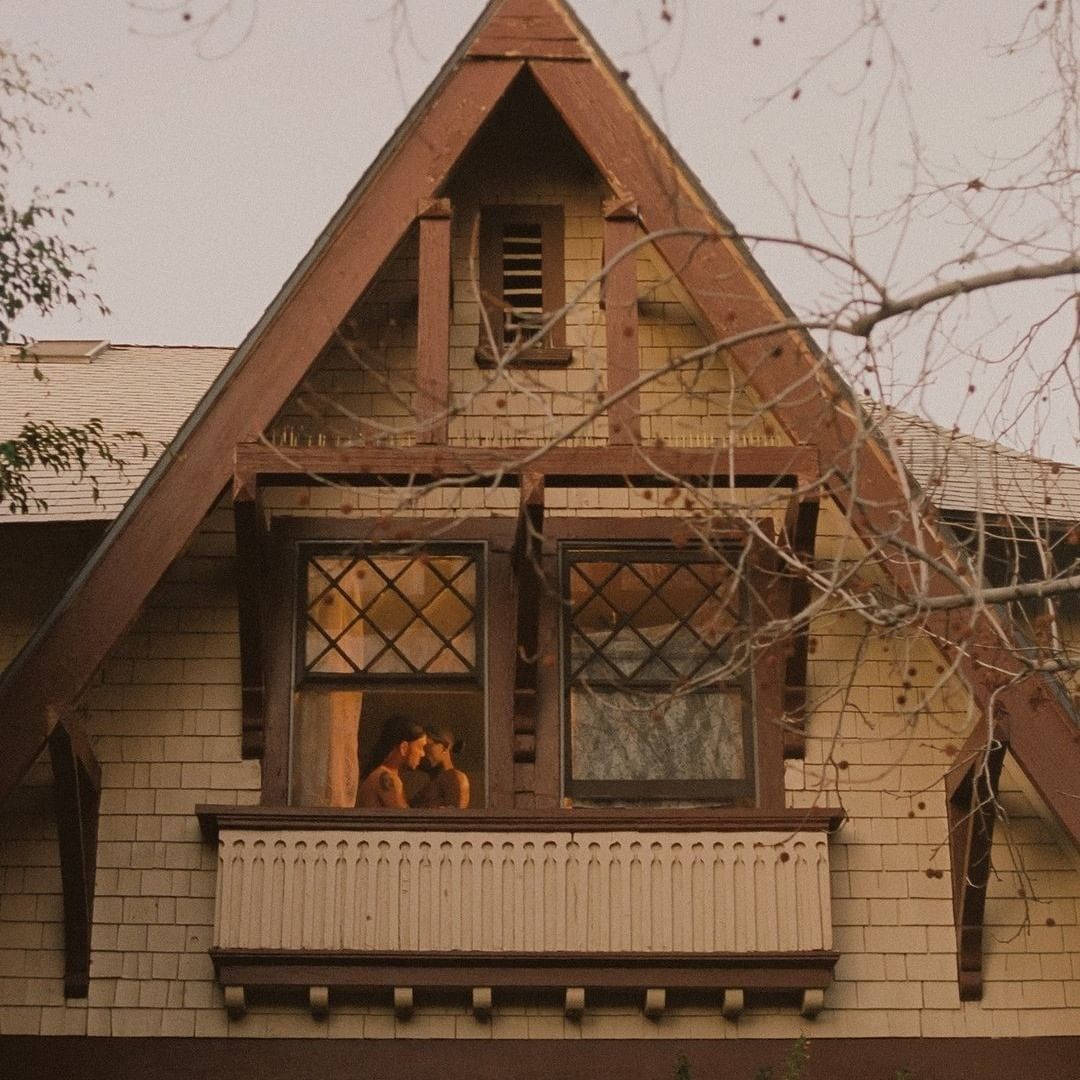 Two people embrace in a house window, set against a backdrop of trees, offering a glimpse into an intimate moment.