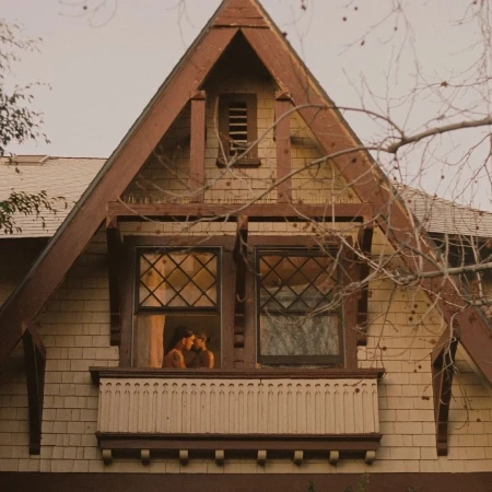 Two people embrace in a house window, set against a backdrop of trees, offering a glimpse into an intimate moment.