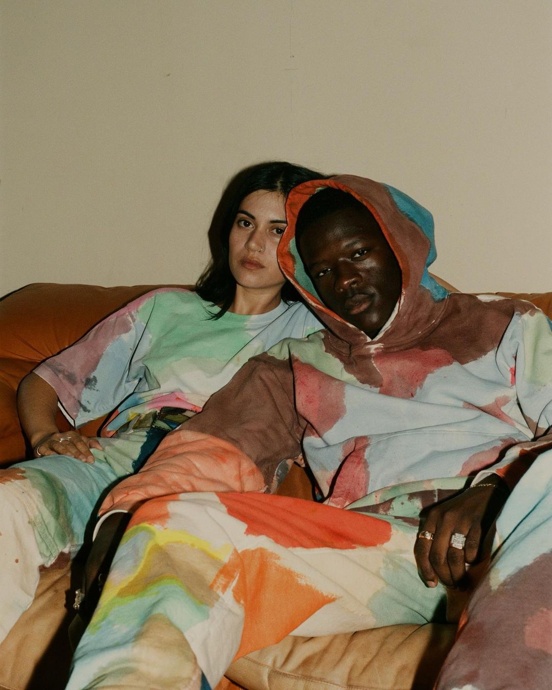 Two people, an adult man and woman, sit on a couch wearing matching watercolor-patterned outfits in an indoor setting, looking at the camera.
