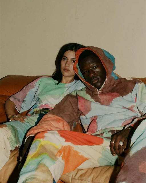 Two people, an adult man and woman, sit on a couch wearing matching watercolor-patterned outfits in an indoor setting, looking at the camera.
