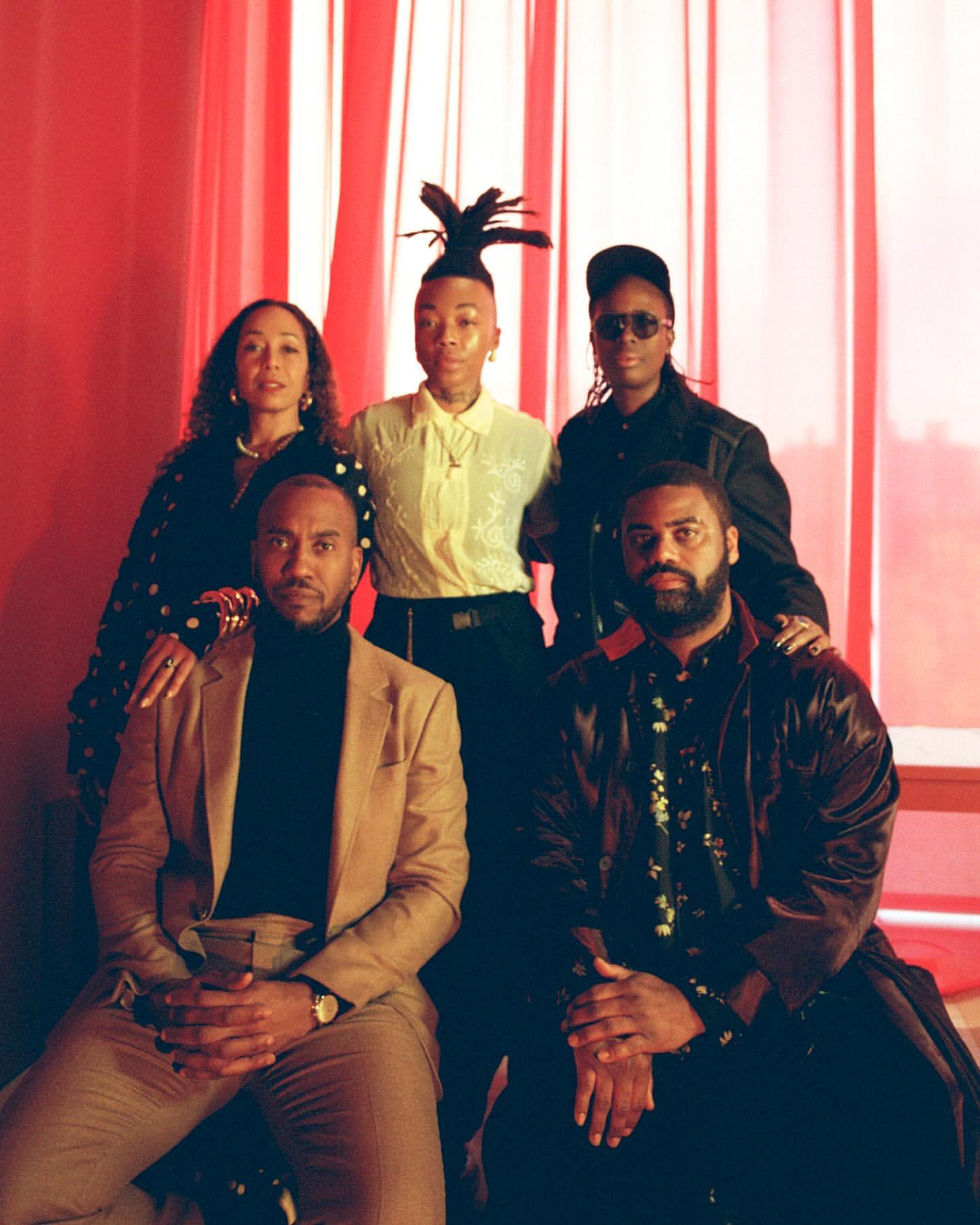 Portrait of five Black individuals posed together against a backdrop of vertical red curtains photographed by Clifford Prince King.
